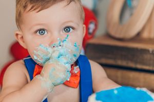 Blue-eyed baby boy with cake-covered hands during a colorful first birthday cake smash photo session.