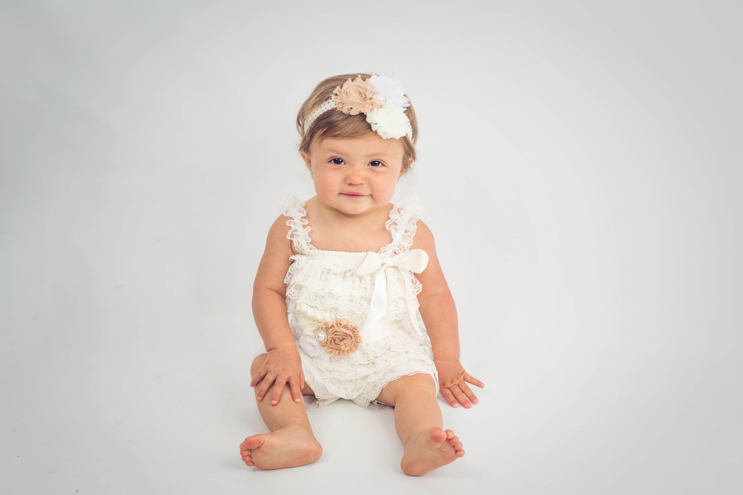 Adorable baby girl in a cream lace romper and floral headband sitting on a white background.