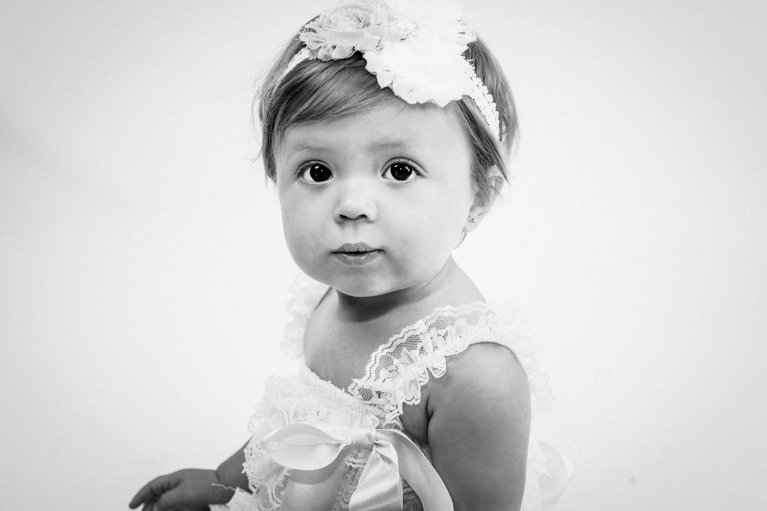 Black and white portrait of a baby girl with a white flower headband wearing a lace dress.