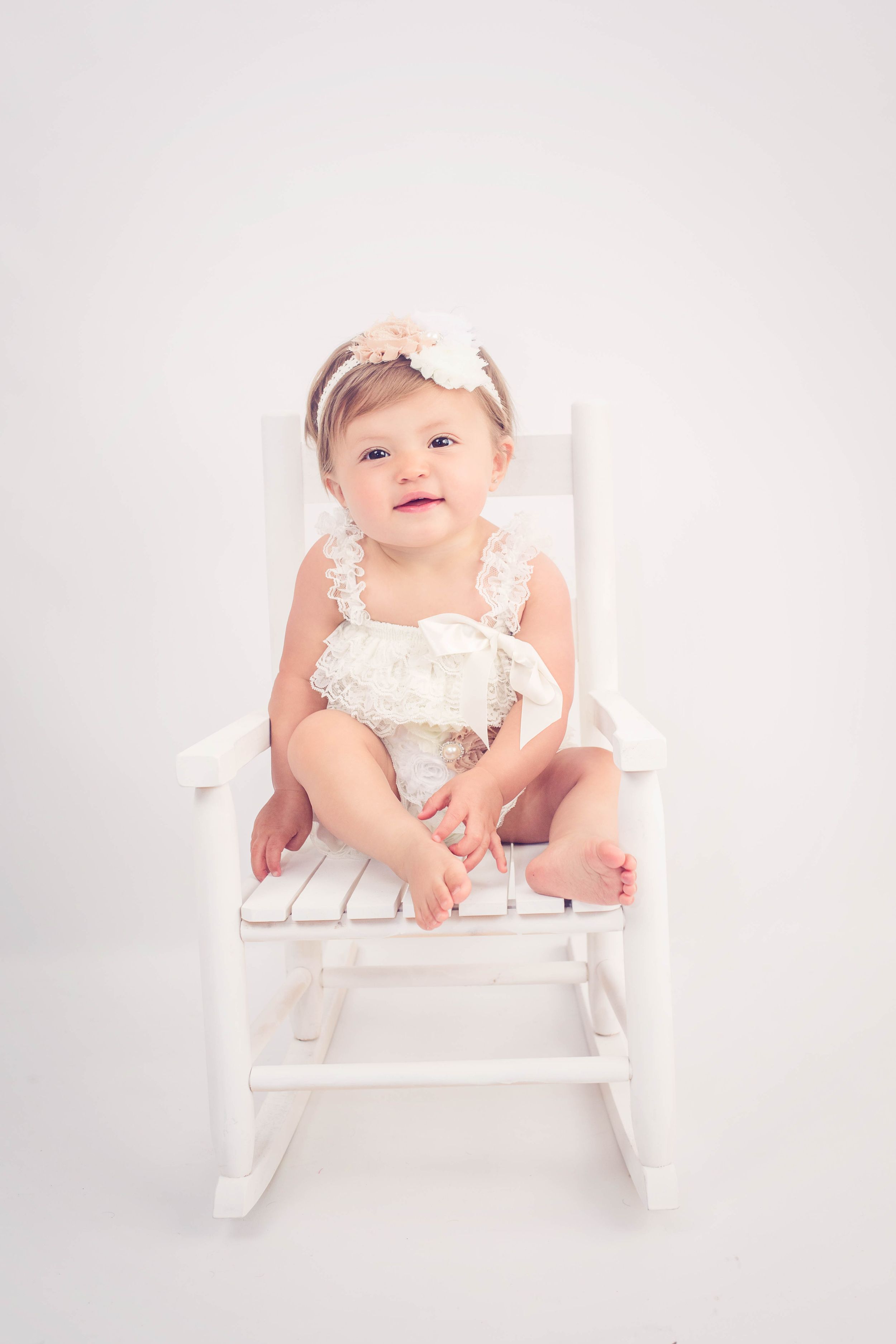 Happy baby girl in a white lace romper and floral headband sitting in a white rocking chair.