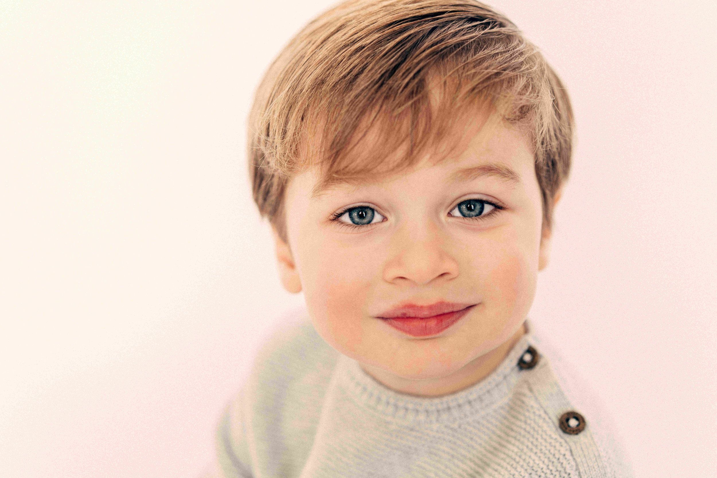 Smiling toddler boy with blue eyes and short blonde hair wearing a gray sweater against a pink background.