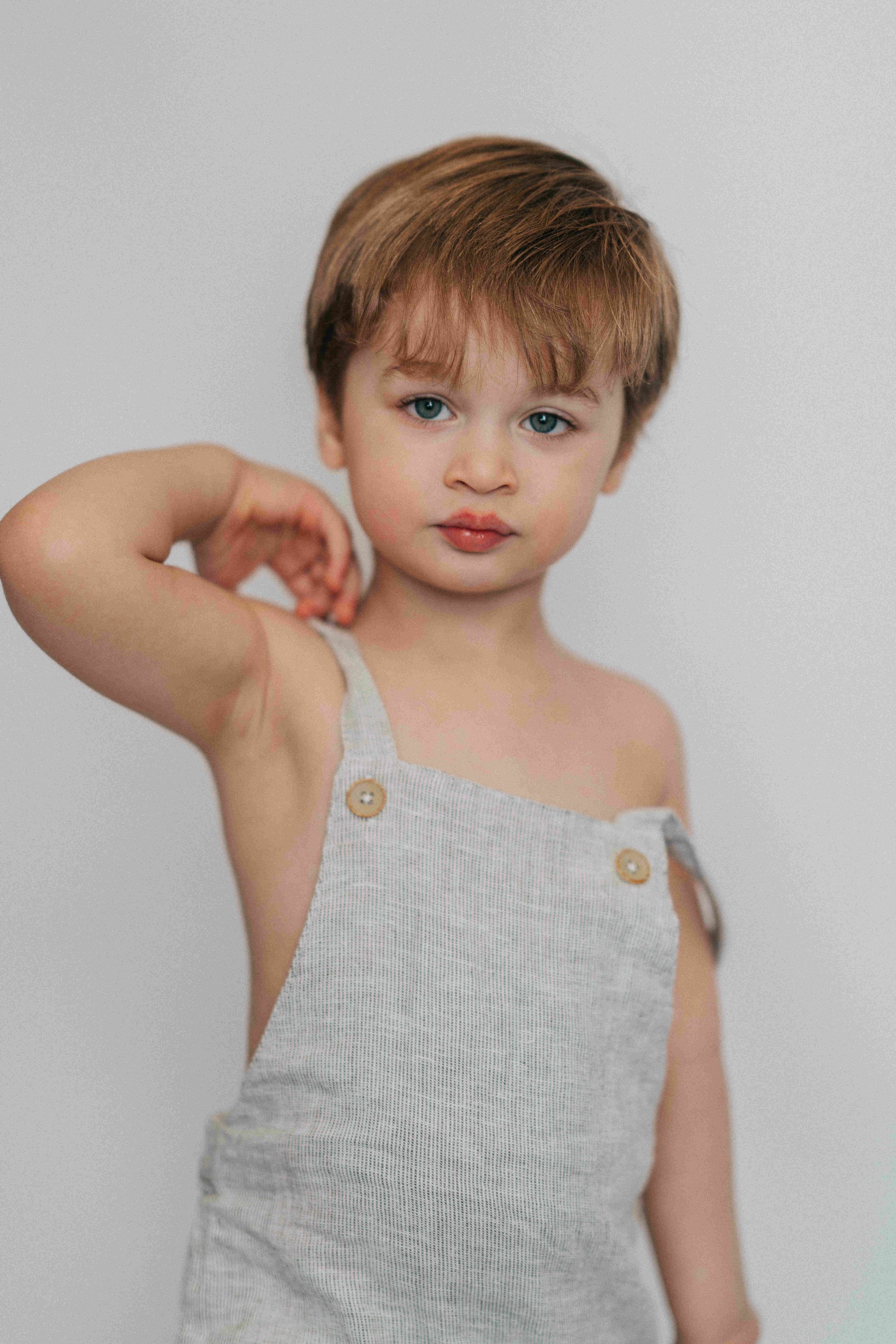 Adorable toddler in gray linen overalls posing against white background with arm raised.