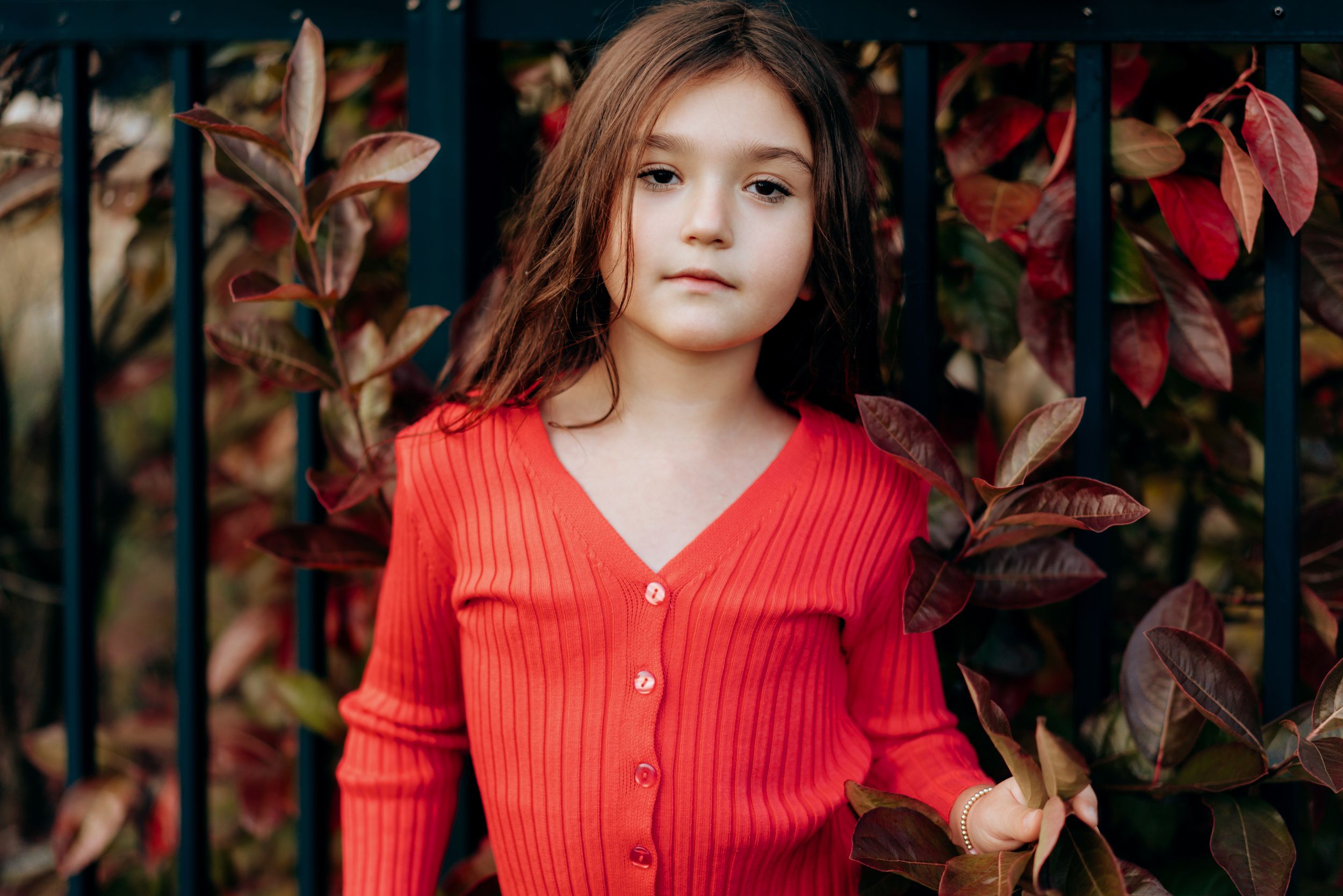 Young girl in red ribbed cardigan standing among colorful autumn leaves by iron fence.