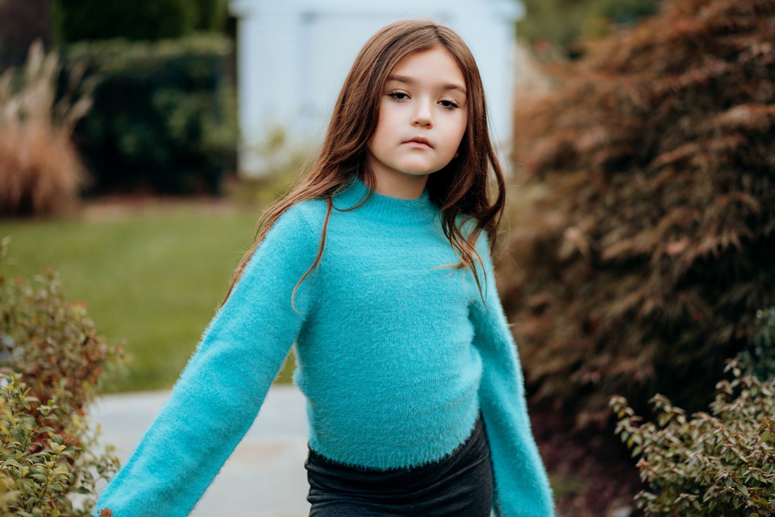 Young girl in teal fuzzy turtleneck sweater walking outdoors among garden greenery.