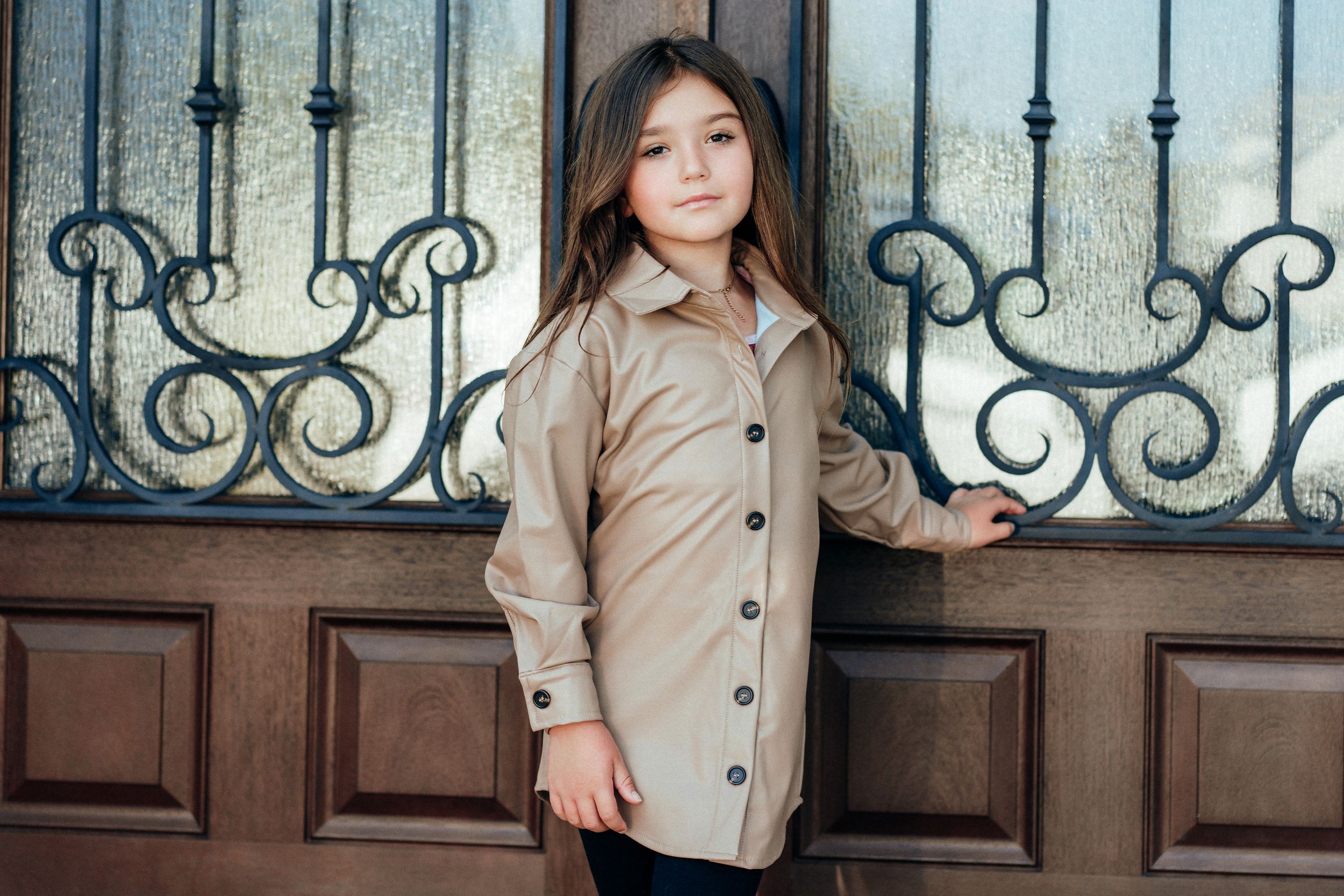 Young girl in beige button-up shirt jacket posing by ornate iron gate and wooden door.