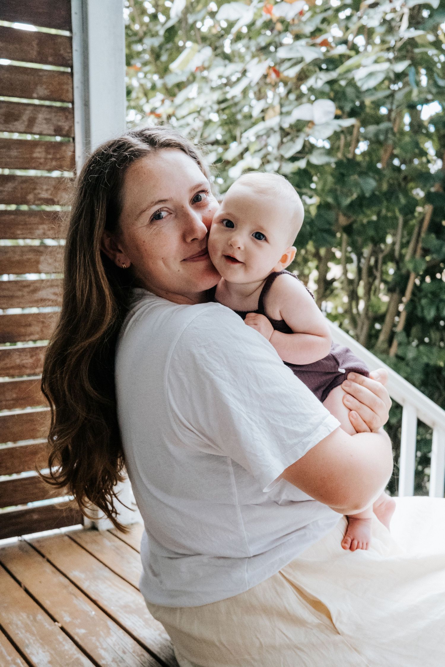 A mother tenderly kisses her baby on a wooden porch, with lush green trees in the background.
