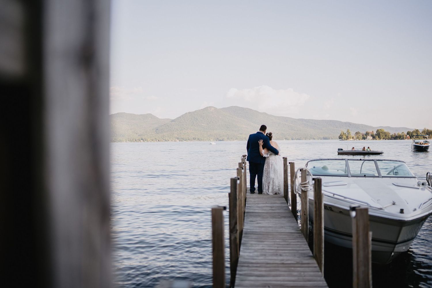 Couple standing on a wooden dock overlooking a serene lake with mountains and a boat nearby.
