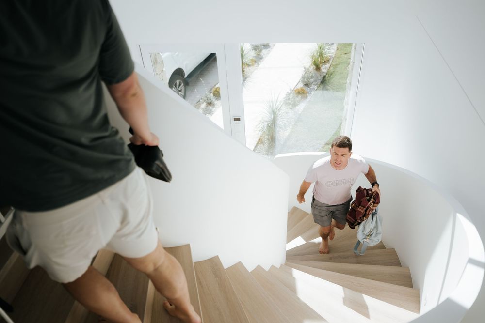 Two men carrying items up modern white interior stairs with wooden steps in a bright contemporary home.