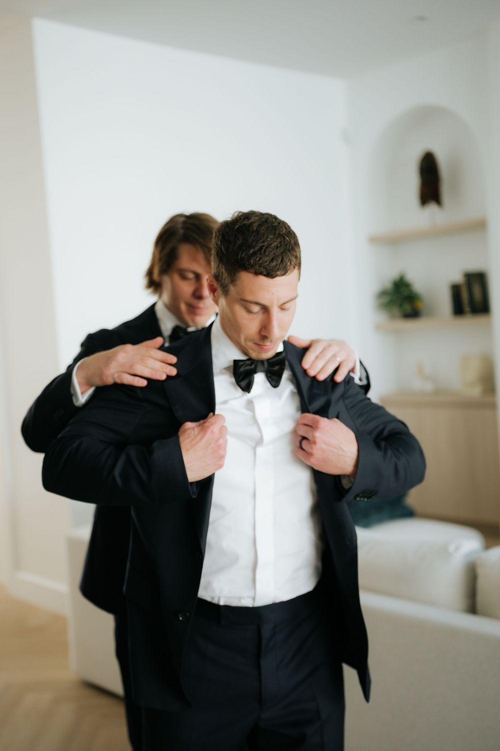 Groom in black tuxedo and bow tie being helped into jacket by groomsman in elegant white interior.