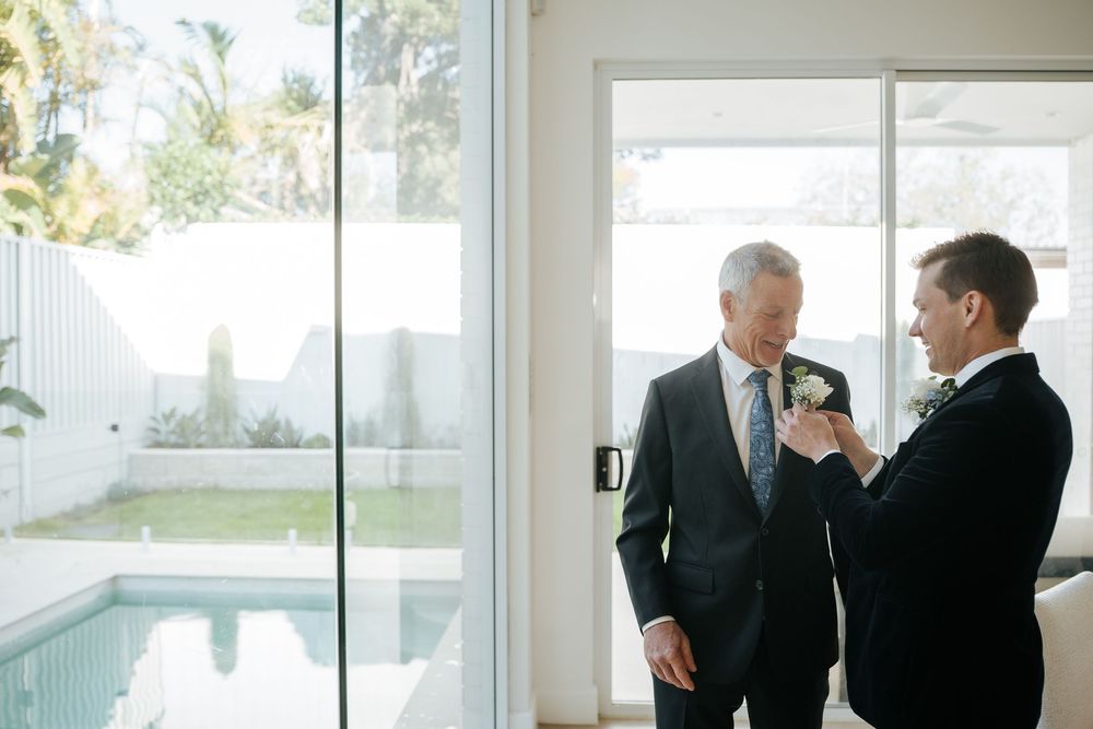Groom adjusting boutonniere on older mans suit jacket indoors near pool at wedding preparation.