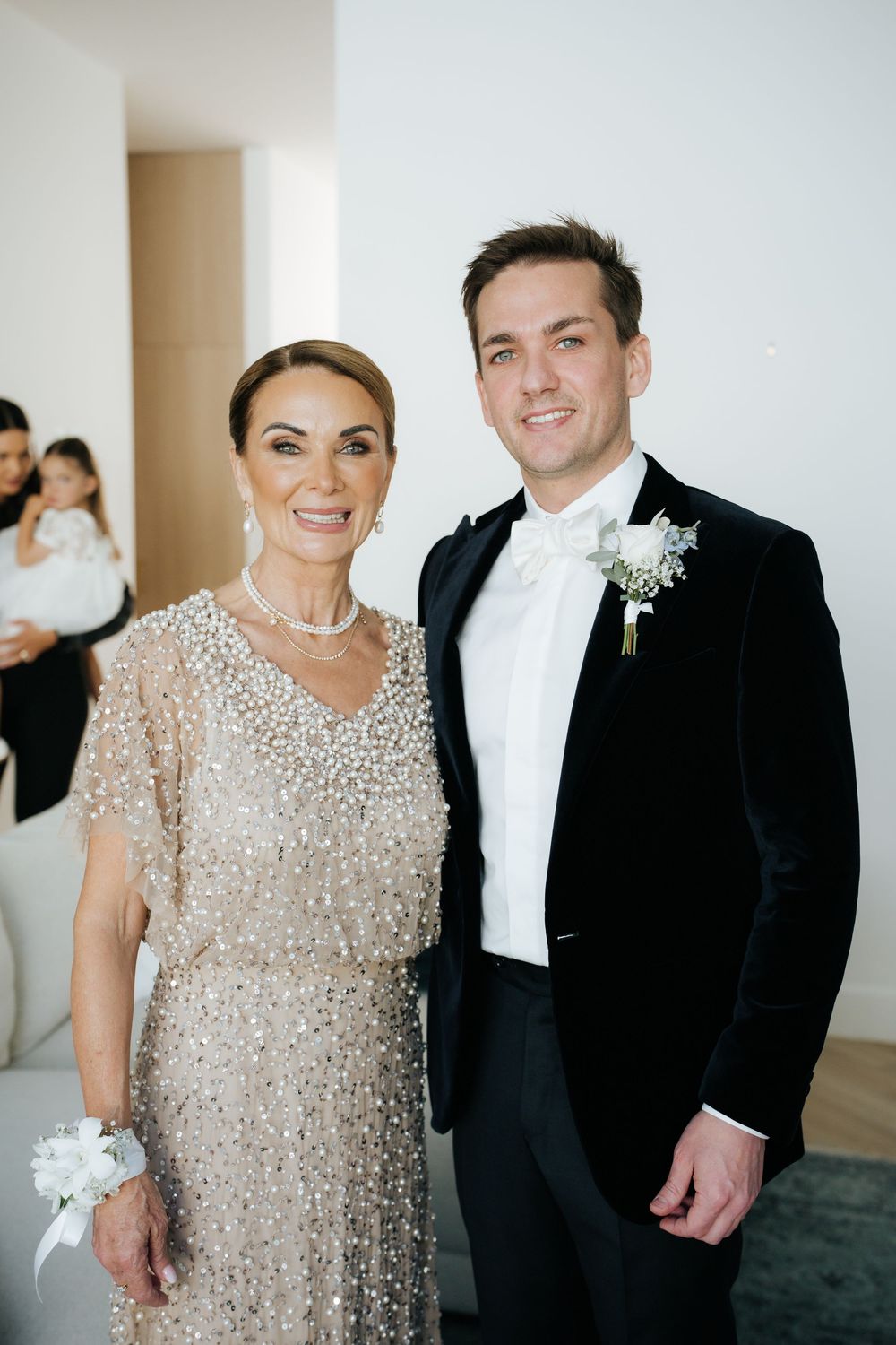Elegant groom in black tuxedo with boutonniere poses with woman in beaded champagne gown at formal wedding event.