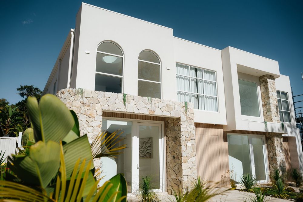 Modern two-story white stucco home with stone accents, arched windows, and tropical landscaping under a clear blue sky.
