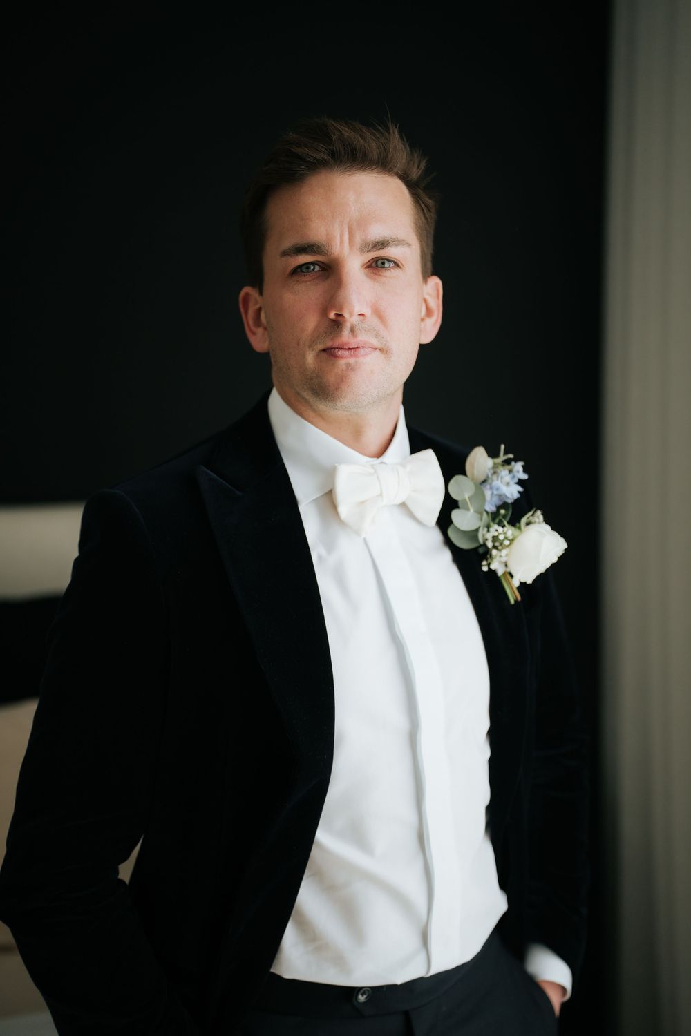 Groom in black velvet tuxedo with white bow tie and floral boutonniere poses confidently for wedding portrait.