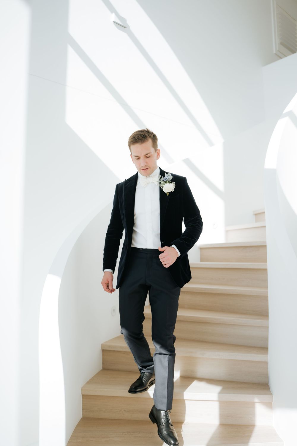 Elegant groom in black tuxedo with boutonniere descending white staircase in bright modern venue.