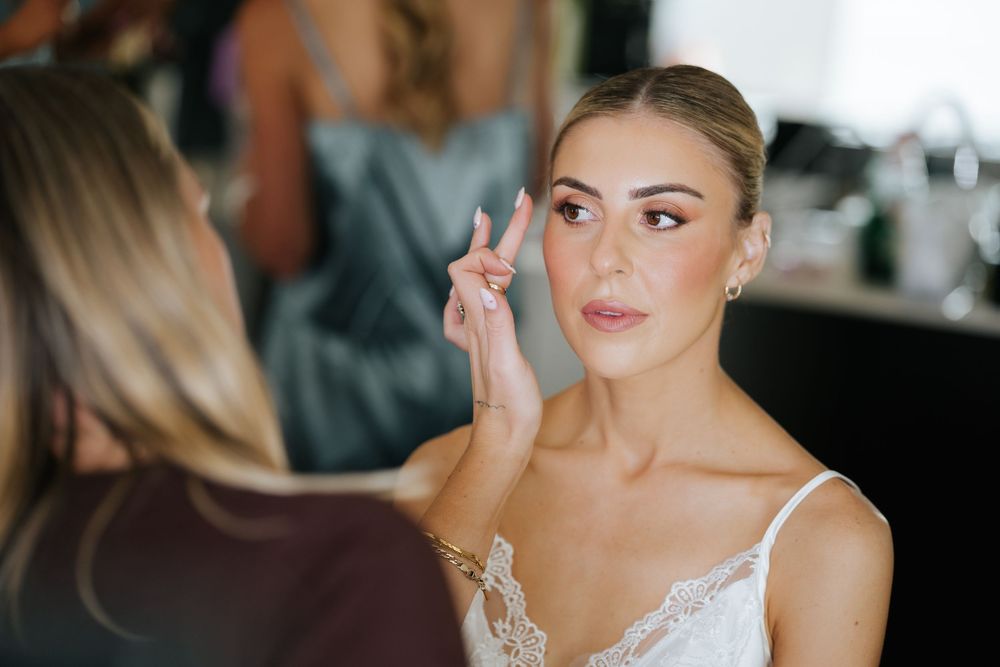 A makeup artist carefully applies eyeshadow to a bride in a white lace gown, preparing her for her wedding day.