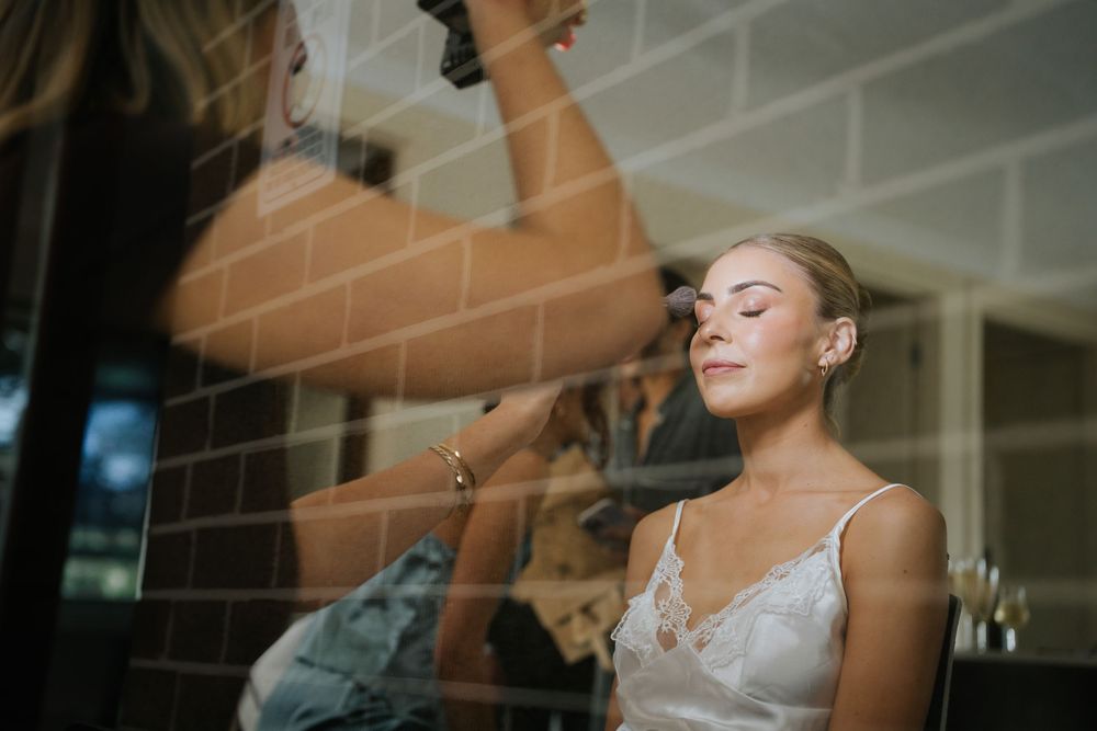 Bride getting makeup applied by artist, eyes closed, wearing white lace top in elegant bridal preparation setting.