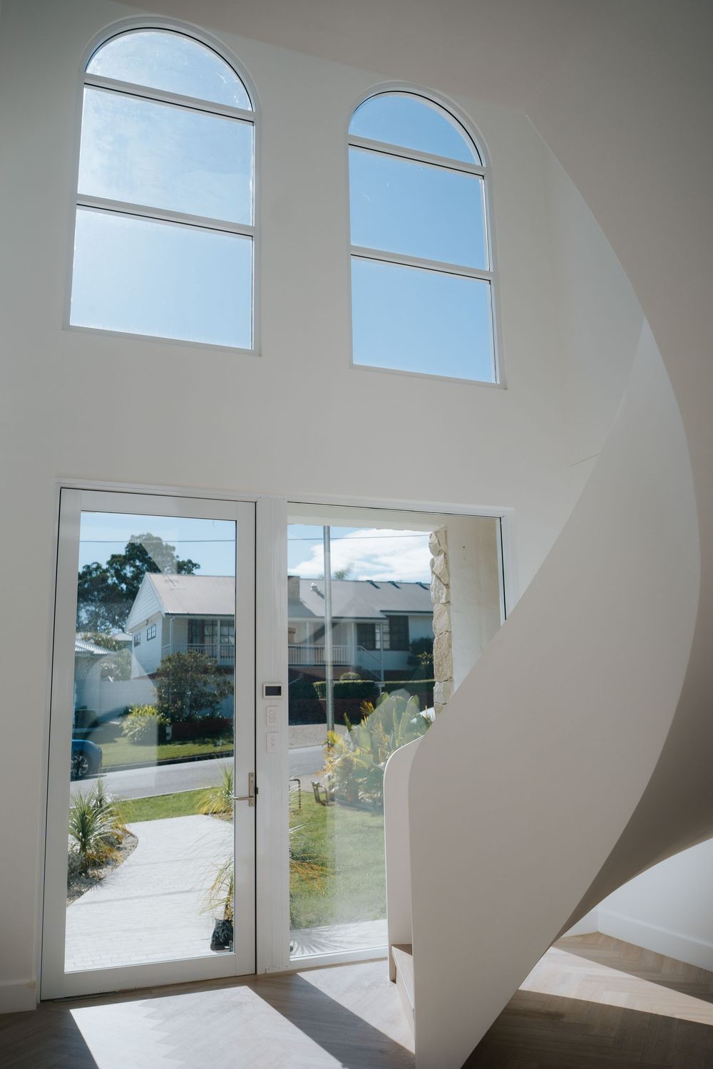Bright white modern entryway with arched windows, glass double doors, and elegant curved staircase flooding space with natural light.