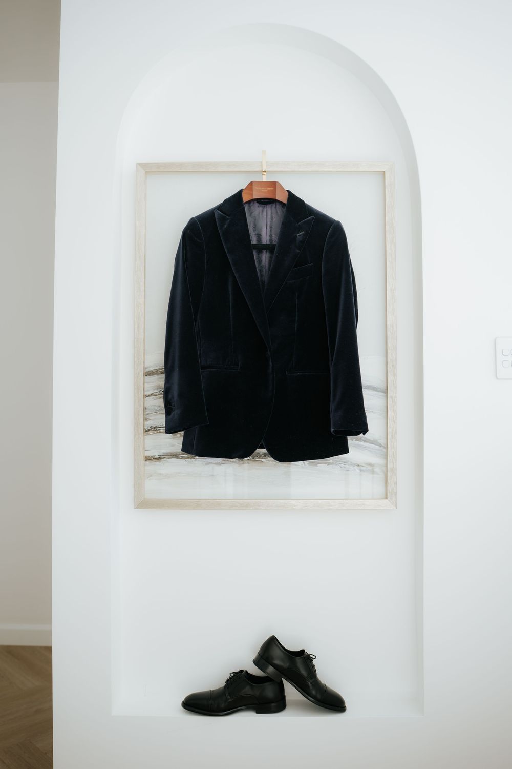 Dark navy velvet blazer on wooden hanger displayed in arched wall niche above black leather dress shoes.