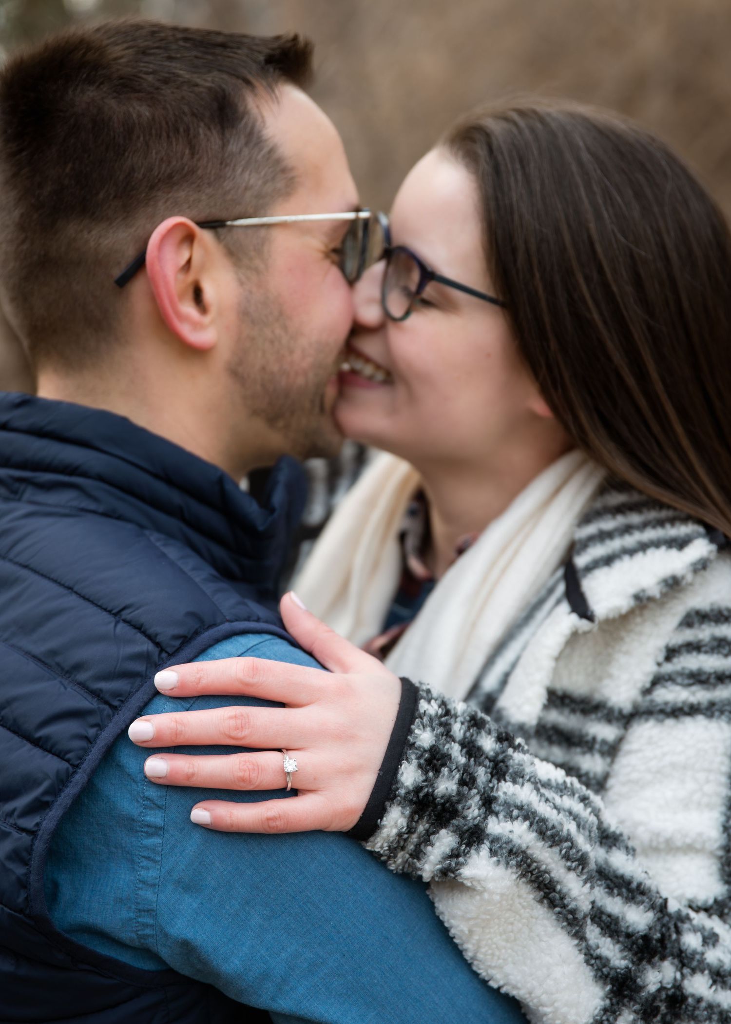 Couple sharing a romantic kiss while embracing outdoors, woman wearing glasses and engagement ring, dressed in winter coats.