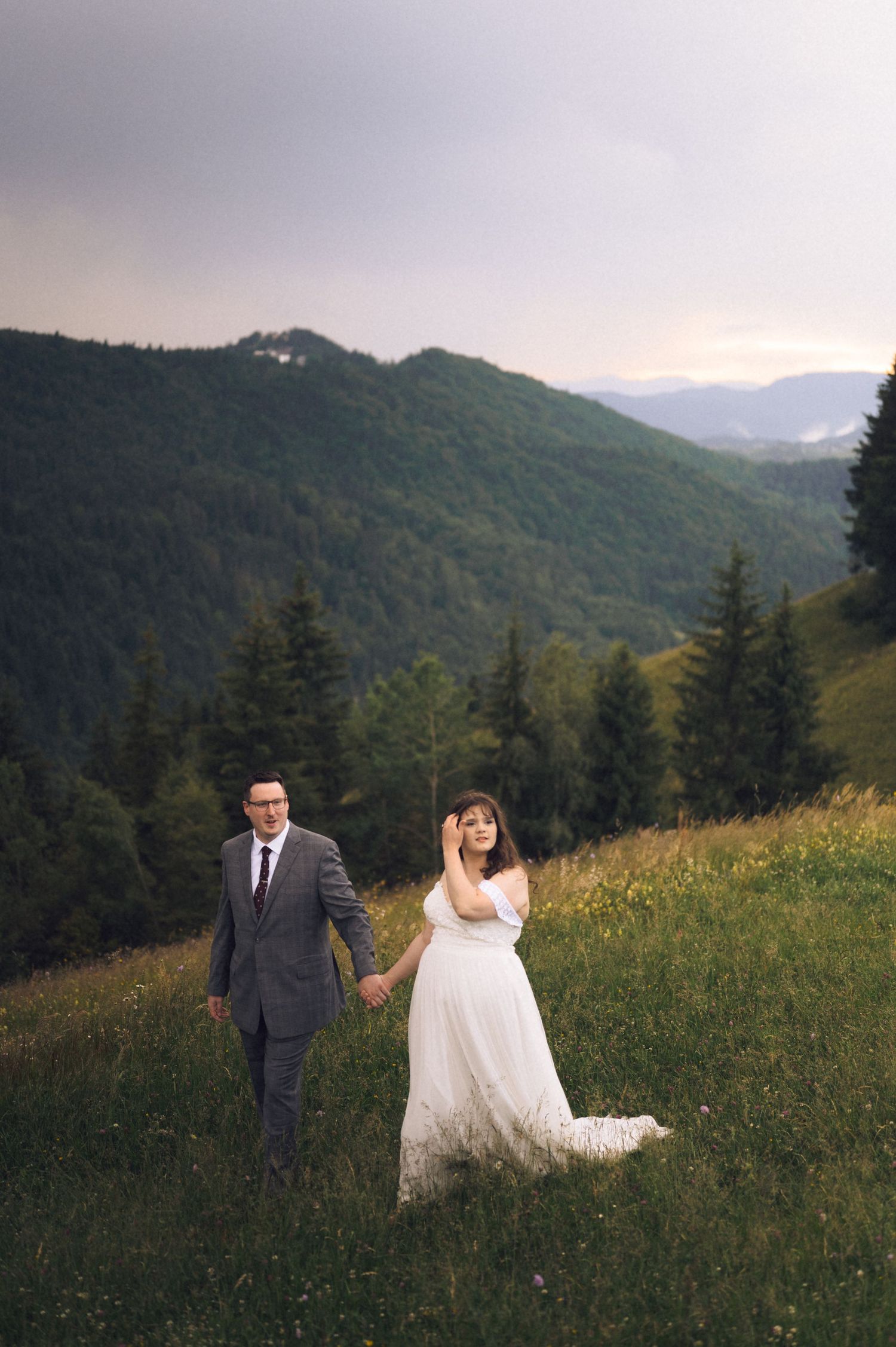 A bride and groom hold hands in a lush mountain meadow at dusk, with forested hills in the background.