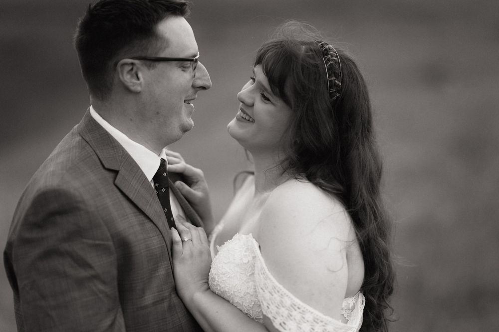 Black and white wedding photo of smiling bride and groom sharing an intimate moment, bride in lace dress with floral headband.