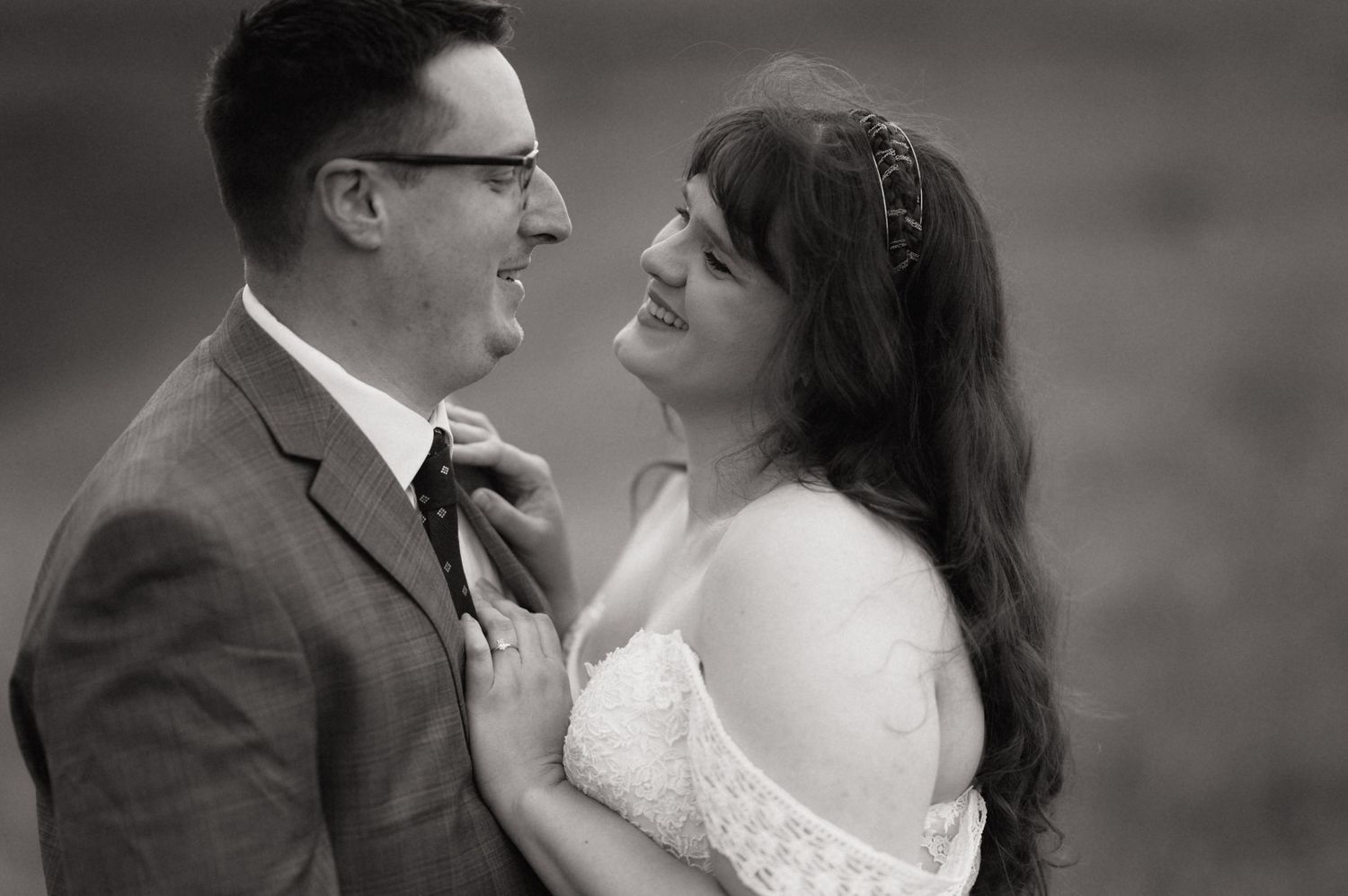 Black and white wedding photo of smiling bride and groom sharing an intimate moment, bride in lace dress with floral headband.