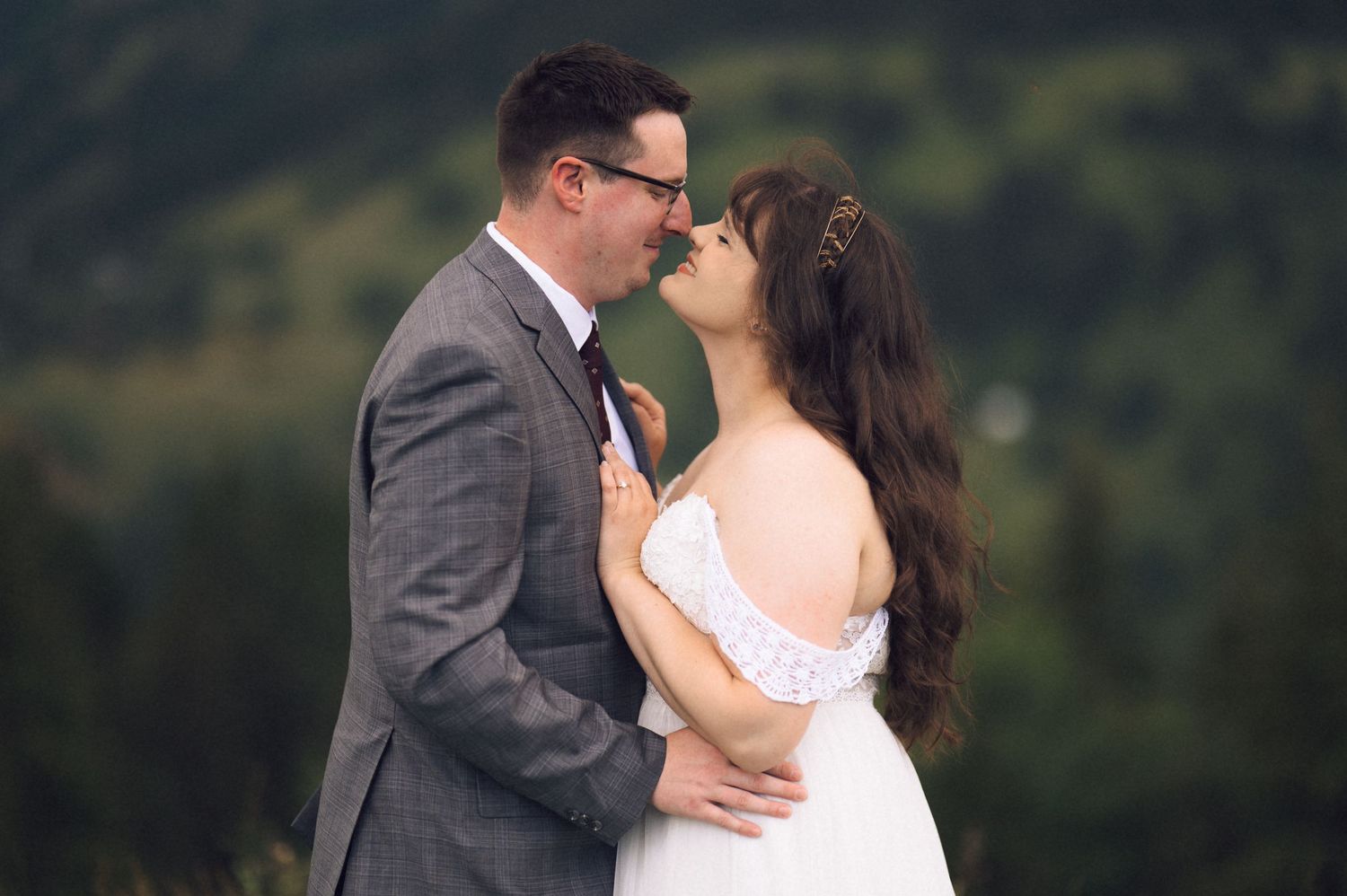 Couple sharing a romantic almost-kiss moment outdoors in wedding attire with lush green forest backdrop.