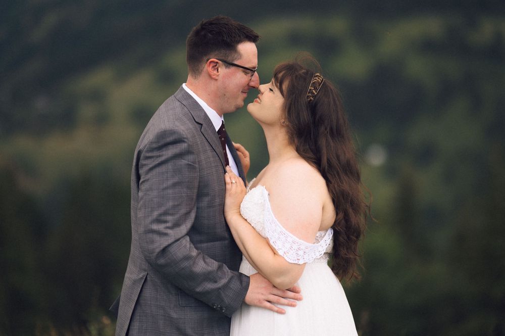 Couple sharing a romantic almost-kiss moment outdoors in wedding attire with lush green forest backdrop.