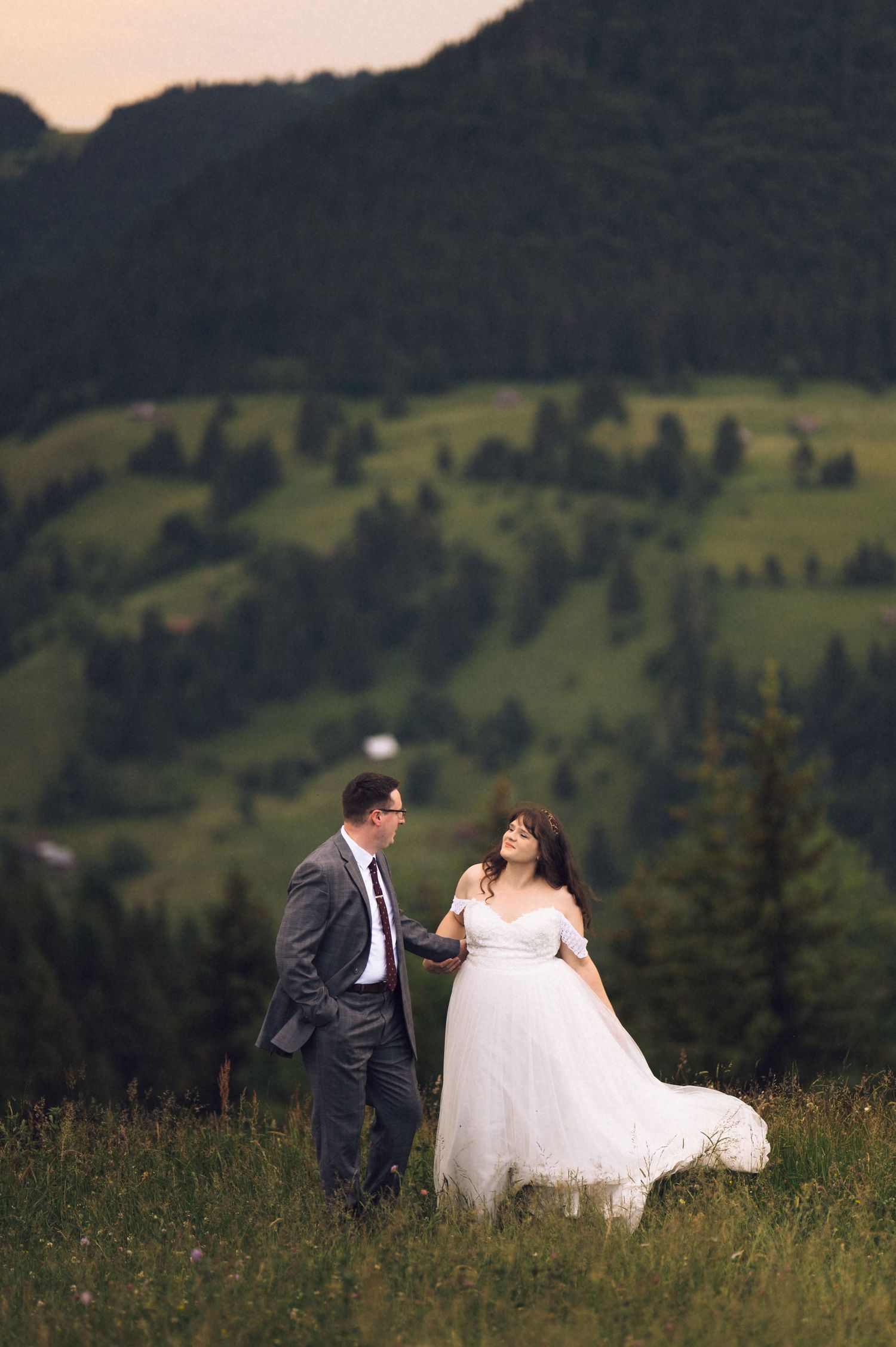 A bride and groom walk hand in hand through a lush green mountain meadow at dusk, her white gown flowing in the breeze.