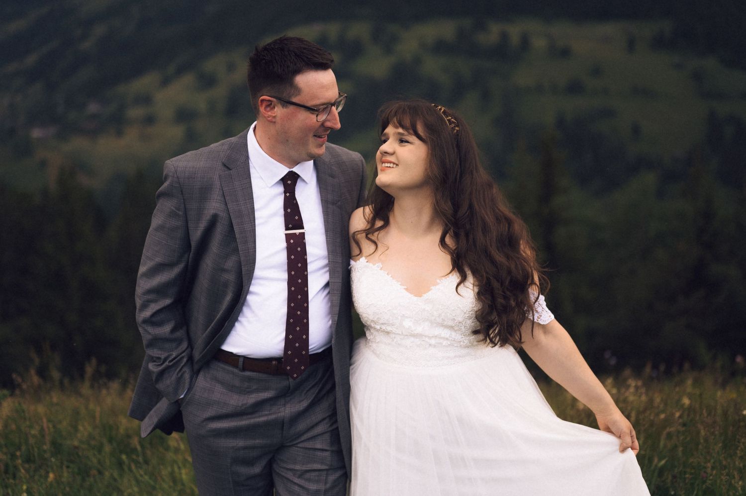 A couple in wedding attire smiles at each other in a lush green mountain meadow, bride in white gown, groom in gray suit.