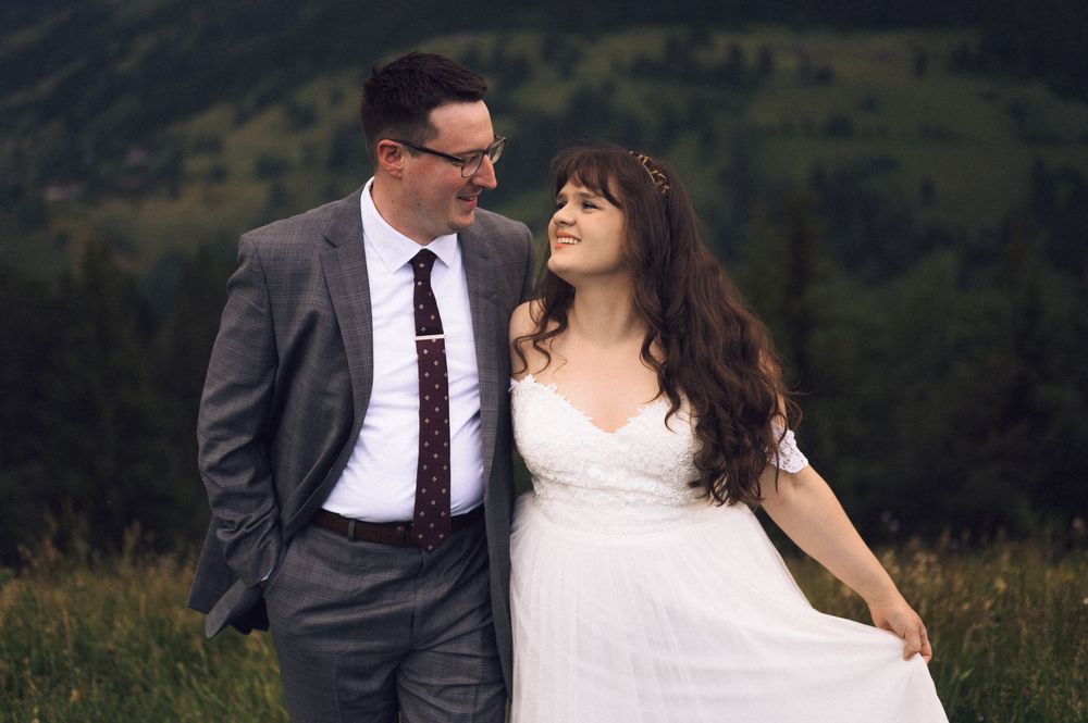 A couple in wedding attire smiles at each other in a lush green mountain meadow, bride in white gown, groom in gray suit.