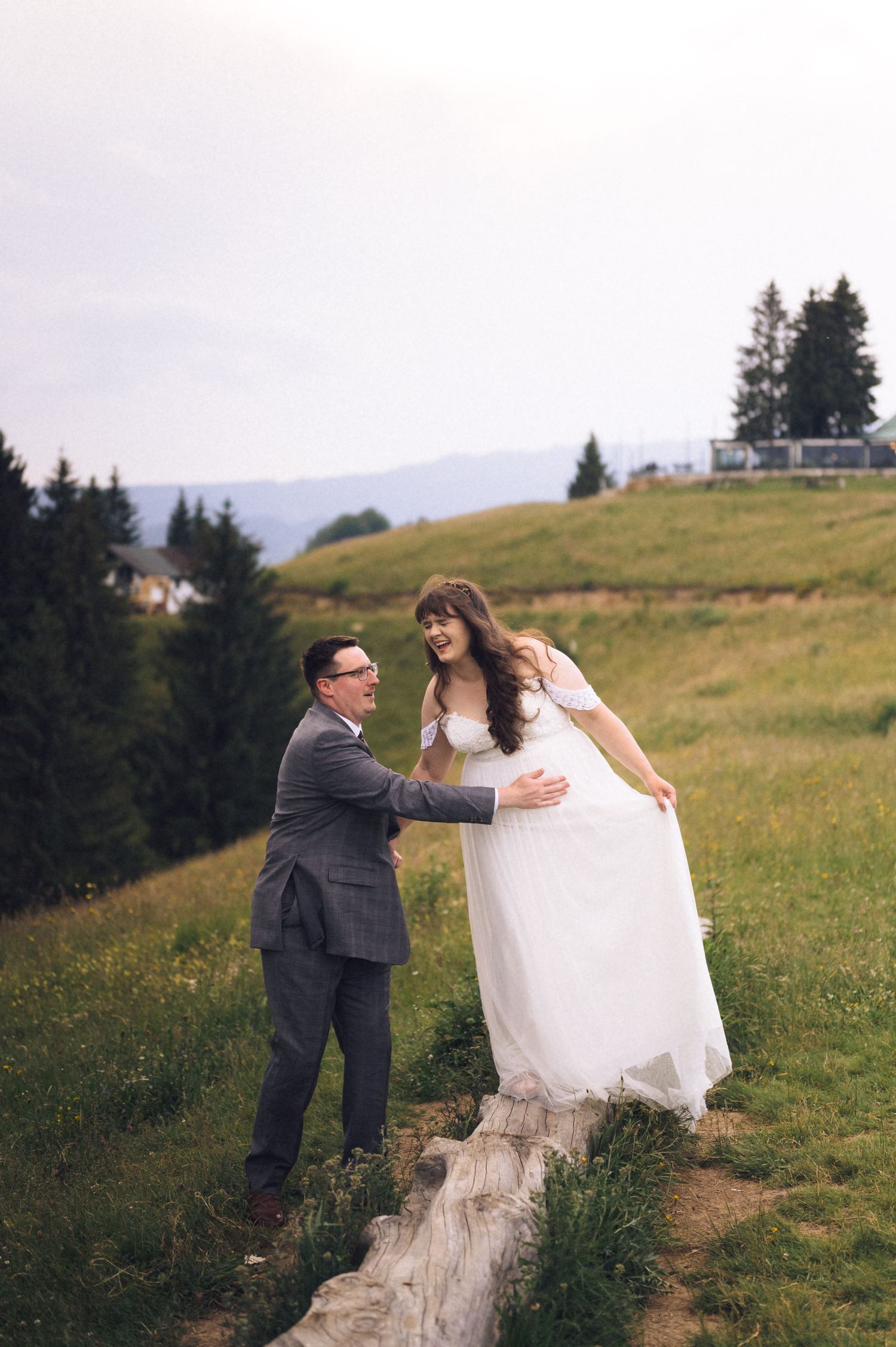 Bride in white gown stands on wooden beam as groom in gray suit steadies her on a lush green hillside.