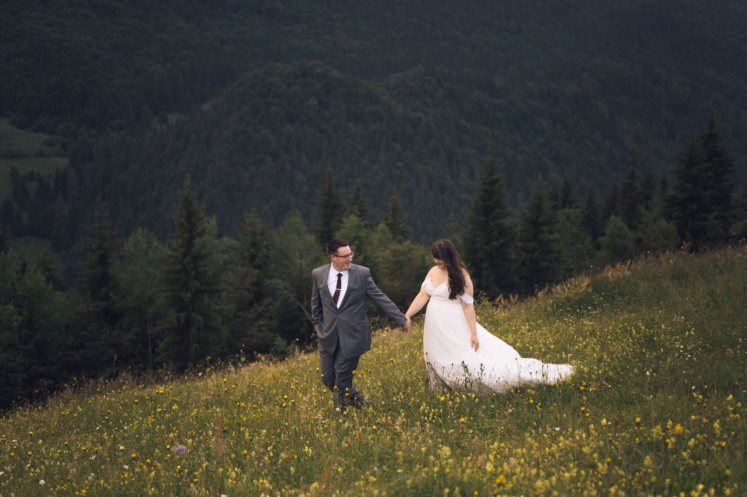 A bride and groom hold hands while walking through a wildflower meadow with lush green forested mountains behind them.