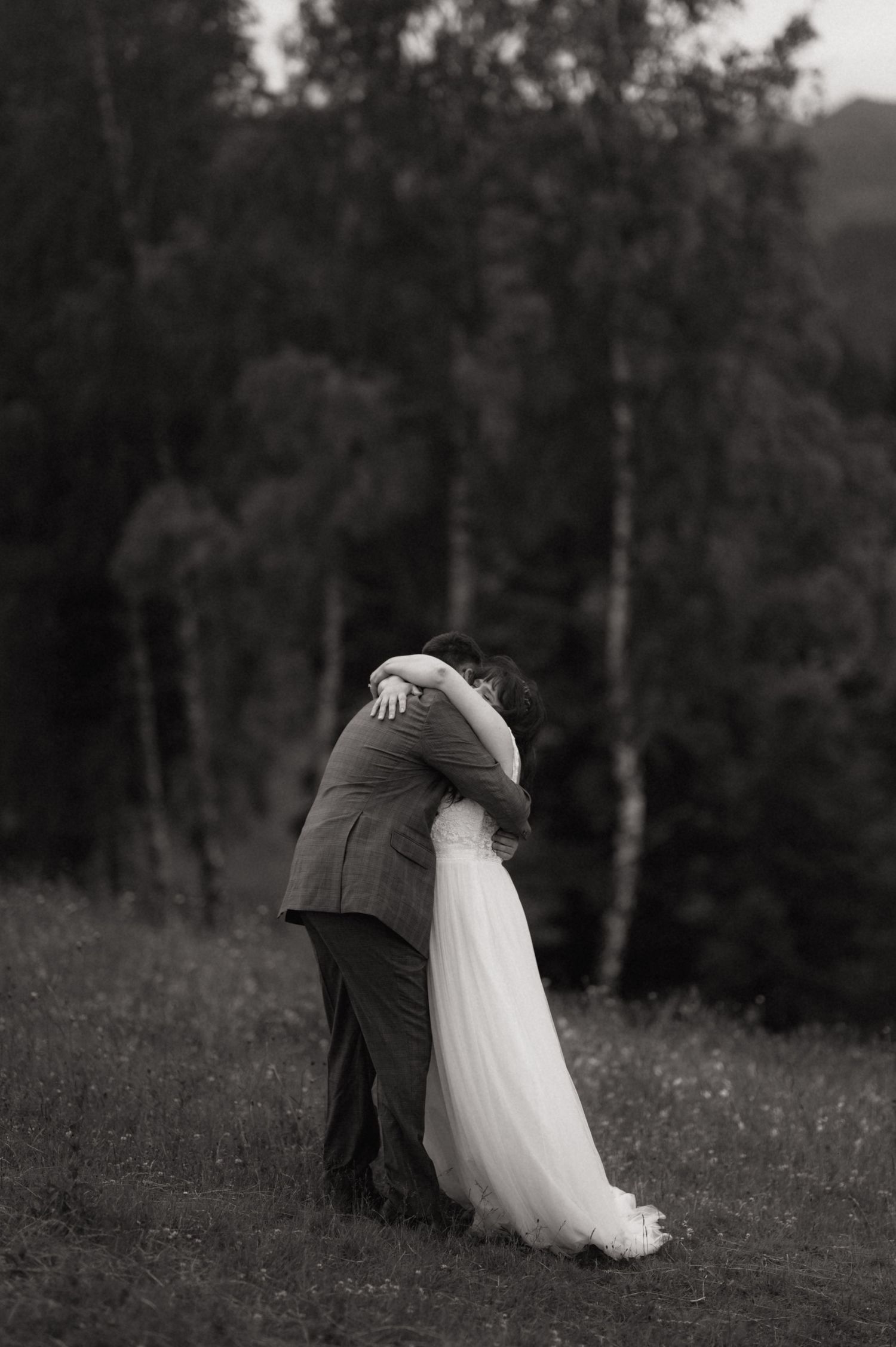 Black and white photo of bride and groom embracing in a field with trees in the background.