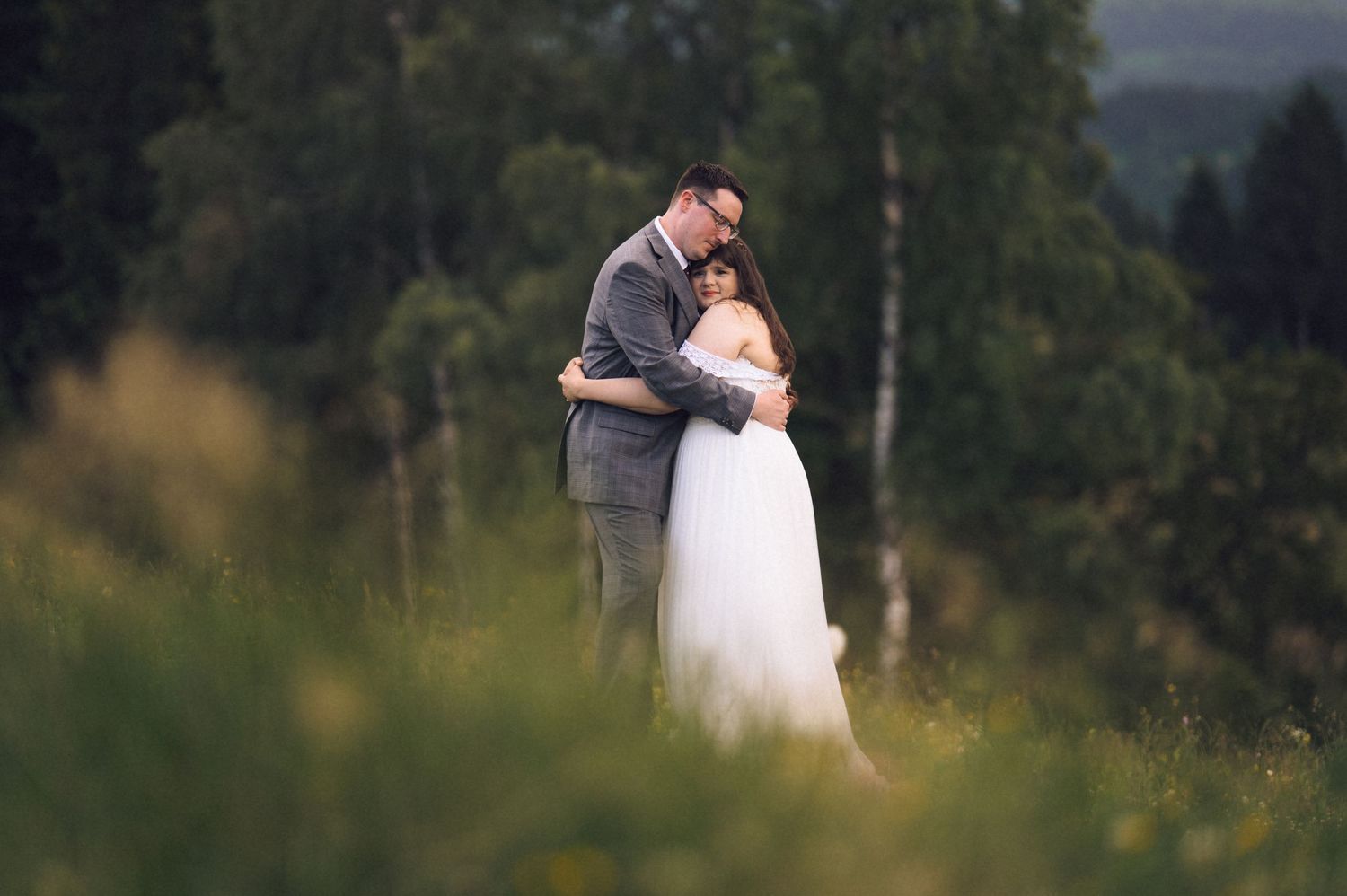 A couple embraces tenderly in a lush green meadow, the bride in white gown and groom in gray suit, surrounded by forest.