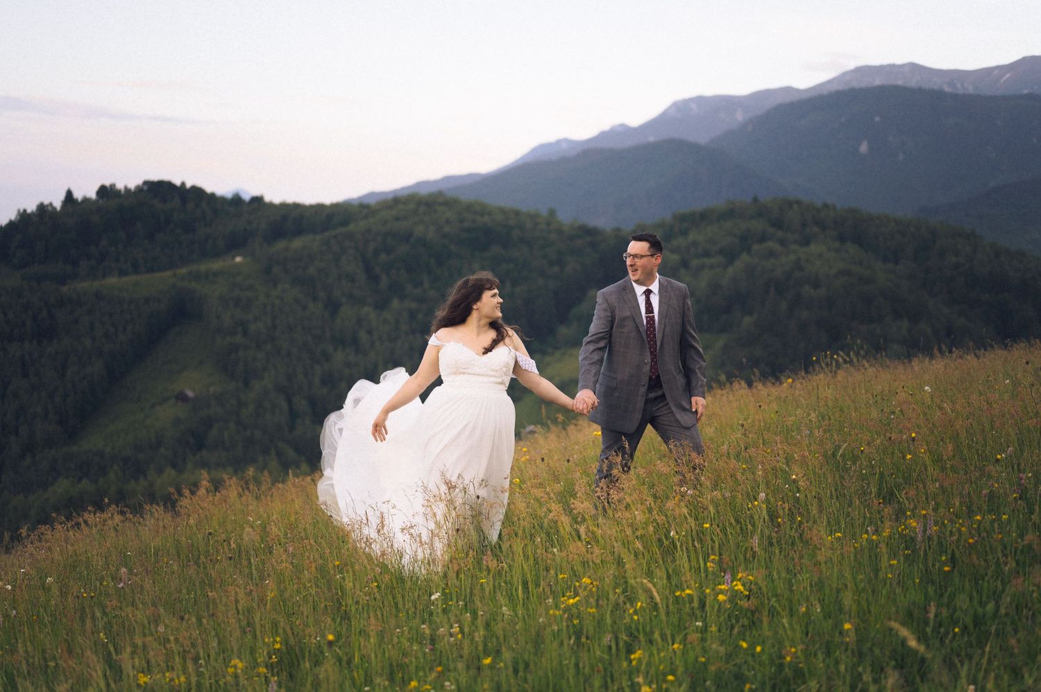 Bride and groom holding hands running through a green mountain meadow in wedding attire.