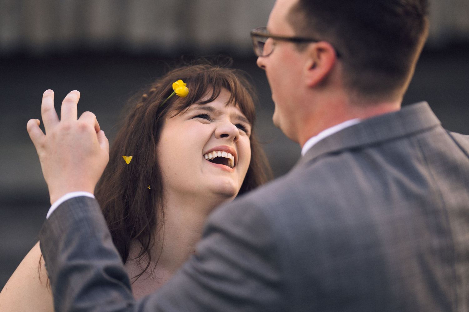 A joyful bride laughs as her groom places yellow flowers in her hair at their outdoor wedding celebration.