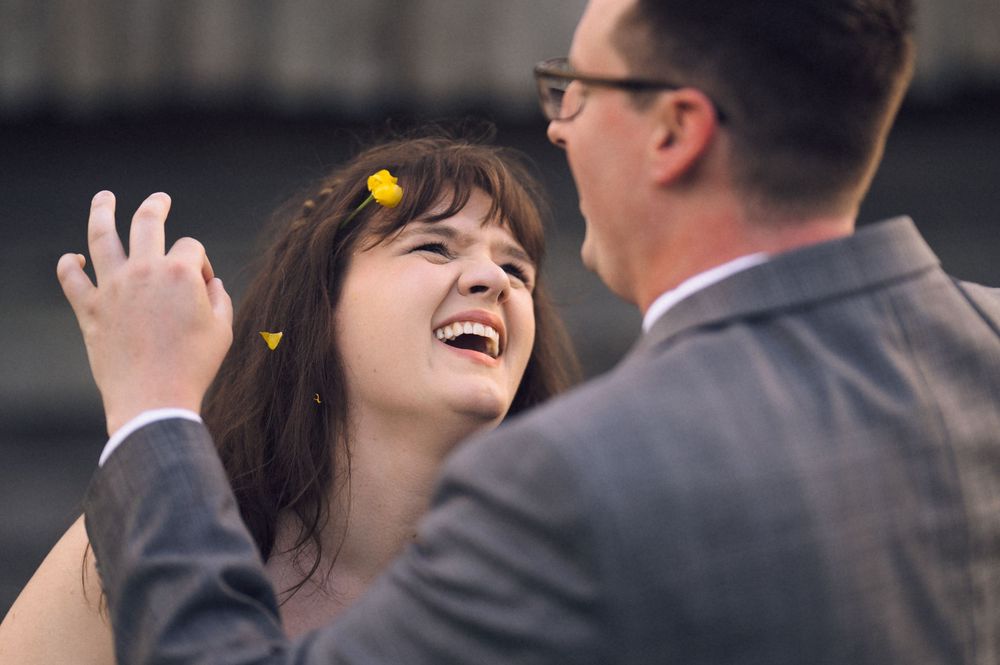 A joyful bride laughs as her groom places yellow flowers in her hair at their outdoor wedding celebration.