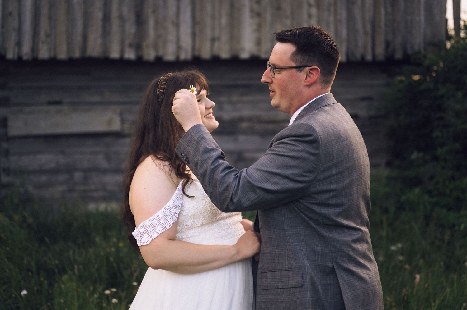 Groom in gray suit gently touches bride's face during outdoor wedding ceremony near rustic barn.