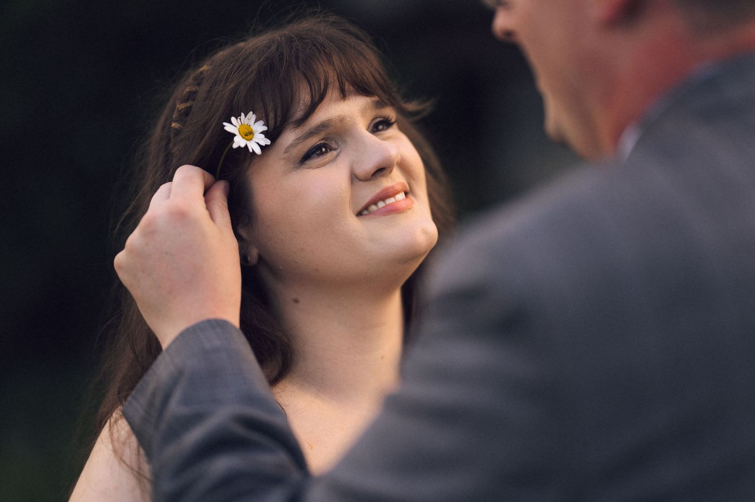 A man gently places a daisy flower in a young woman's dark hair as she smiles up at him outdoors.