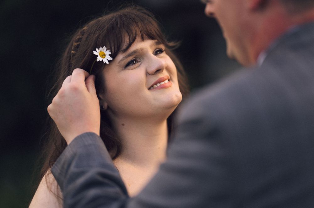 A man gently places a daisy flower in a young woman's dark hair as she smiles up at him outdoors.