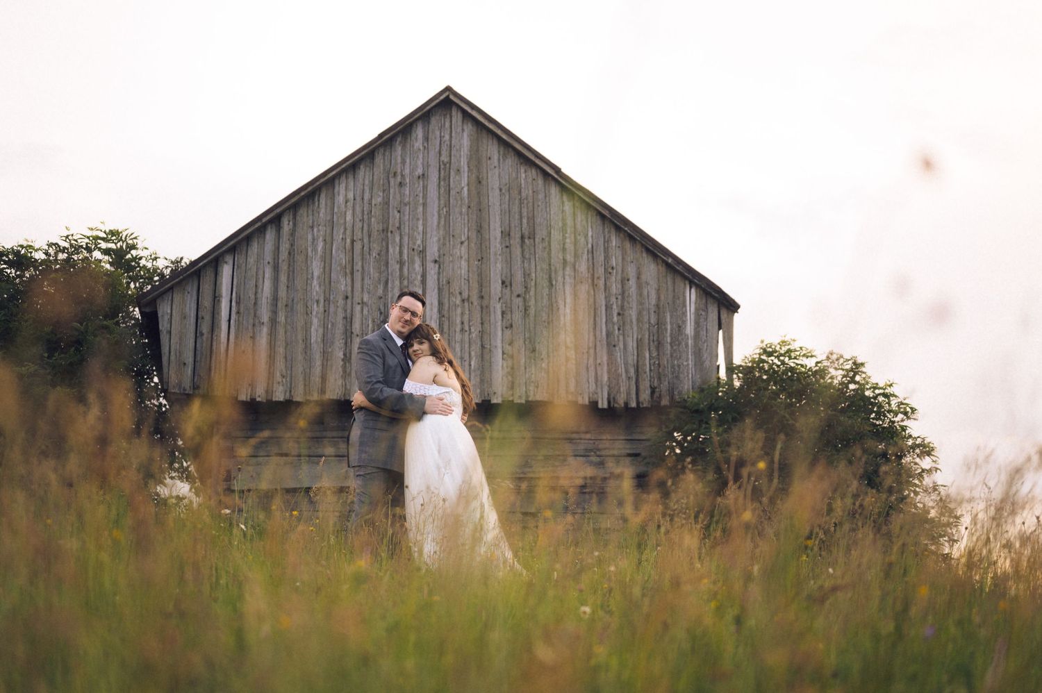 A couple in wedding attire embraces in a field of tall grass before a rustic wooden barn at dusk.