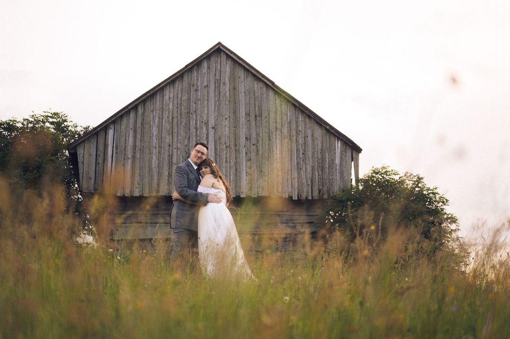 A couple in wedding attire embraces in a field of tall grass before a rustic wooden barn at dusk.