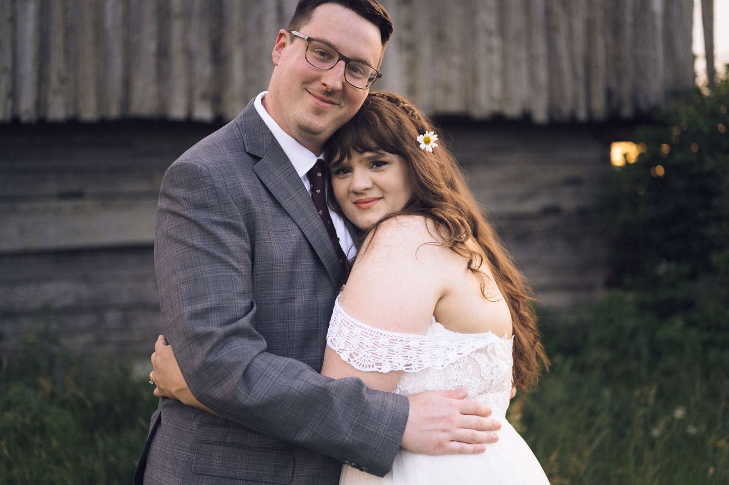 A couple in wedding attire embracing outdoors, groom in gray suit and bride in white lace dress.