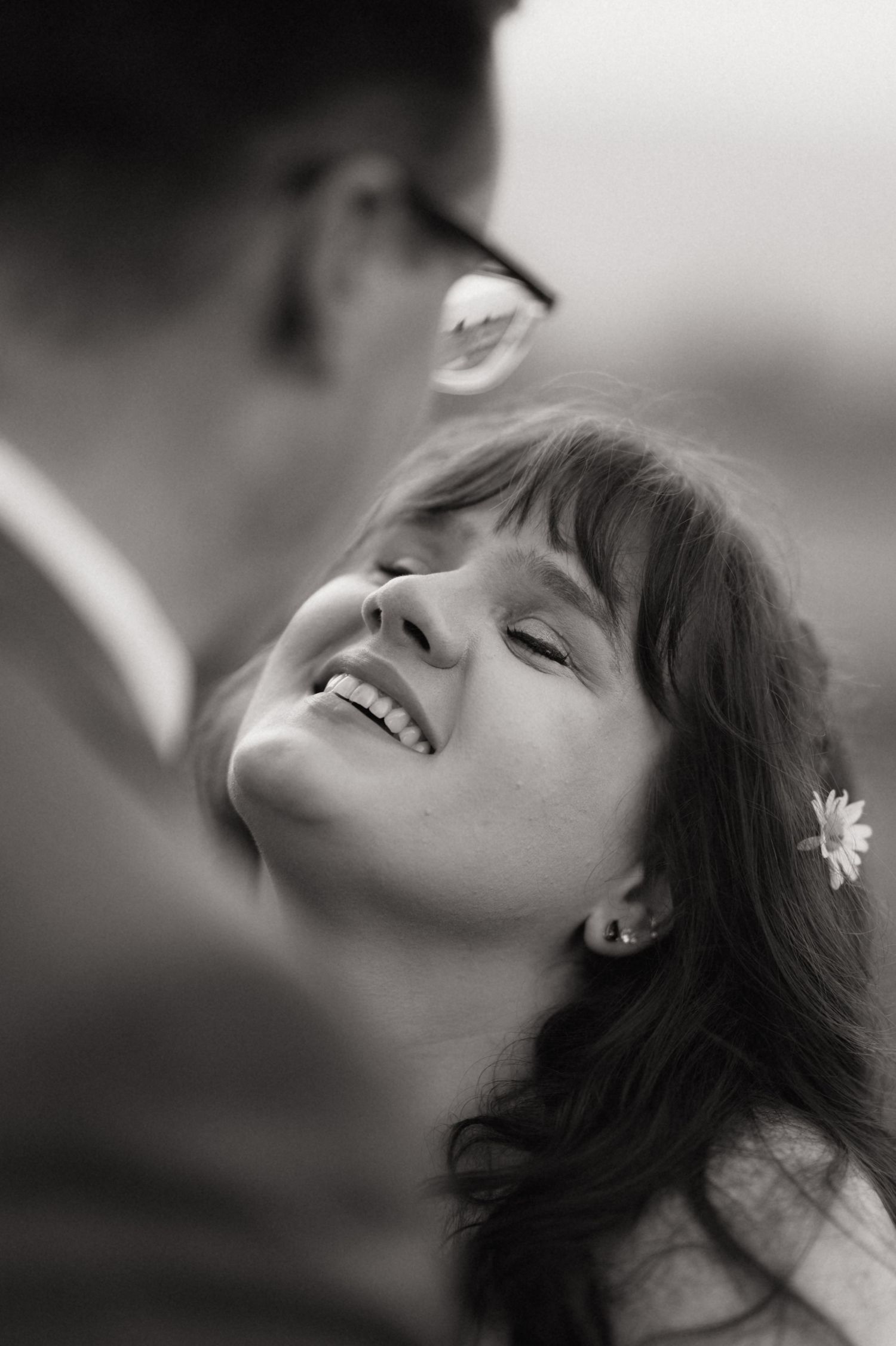 Black and white photo of a smiling woman with a flower in her hair gazing up at a man wearing glasses.
