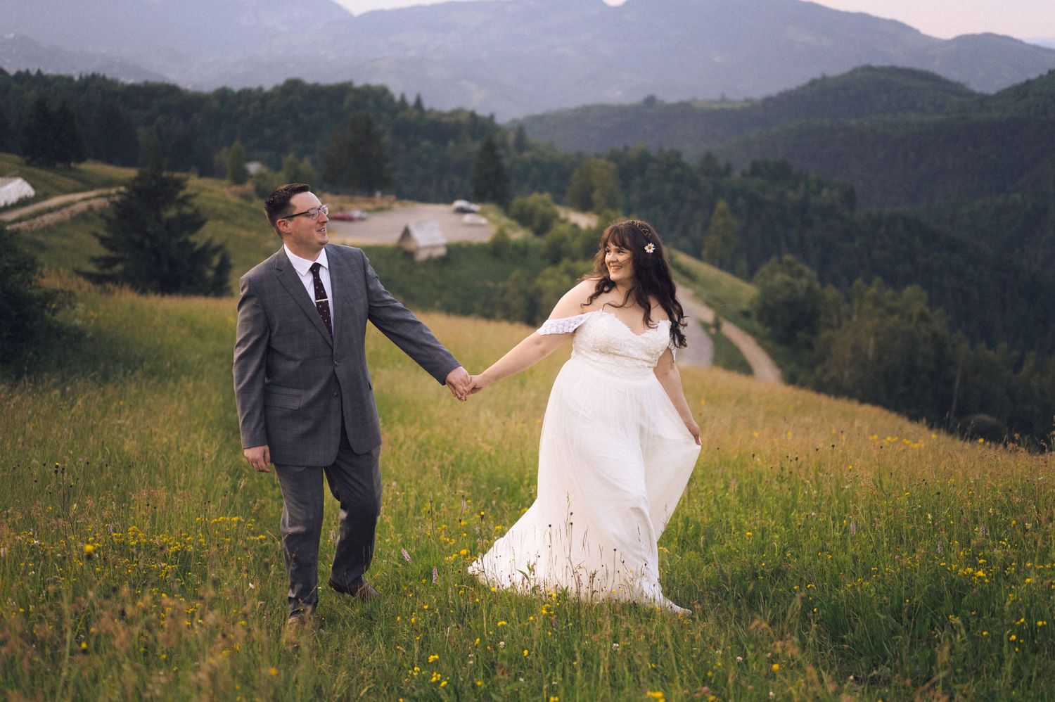 A bride and groom hold hands in a lush green mountain meadow, with scenic hills and trees in the background.