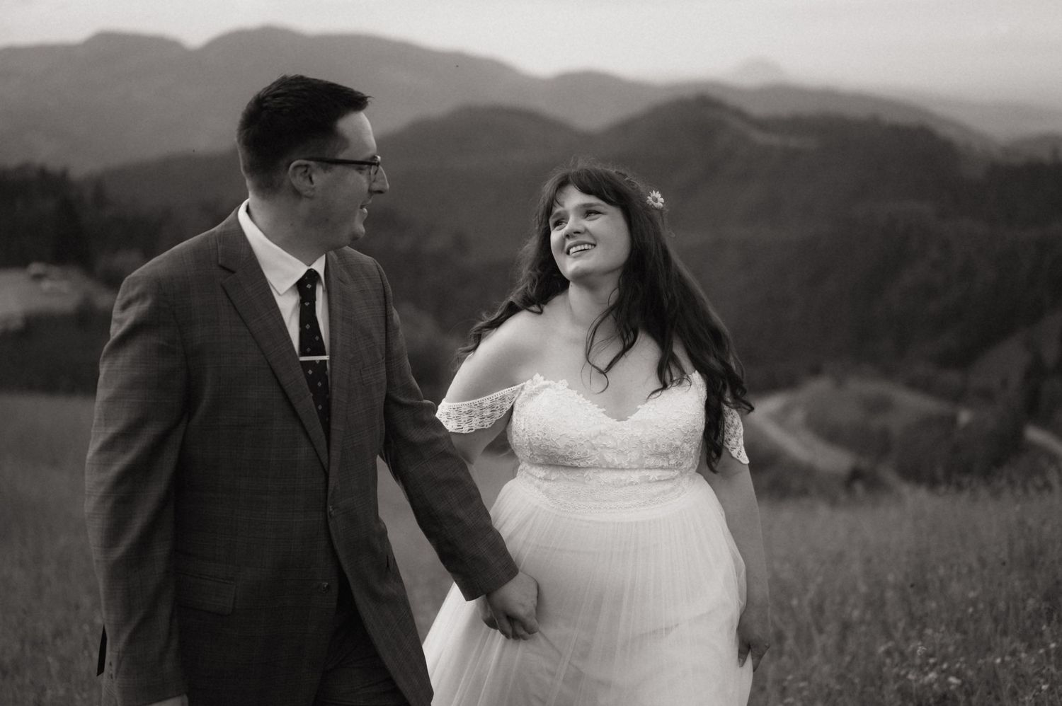 Black and white photo of a bride and groom holding hands in a mountain meadow, sharing a joyful moment.