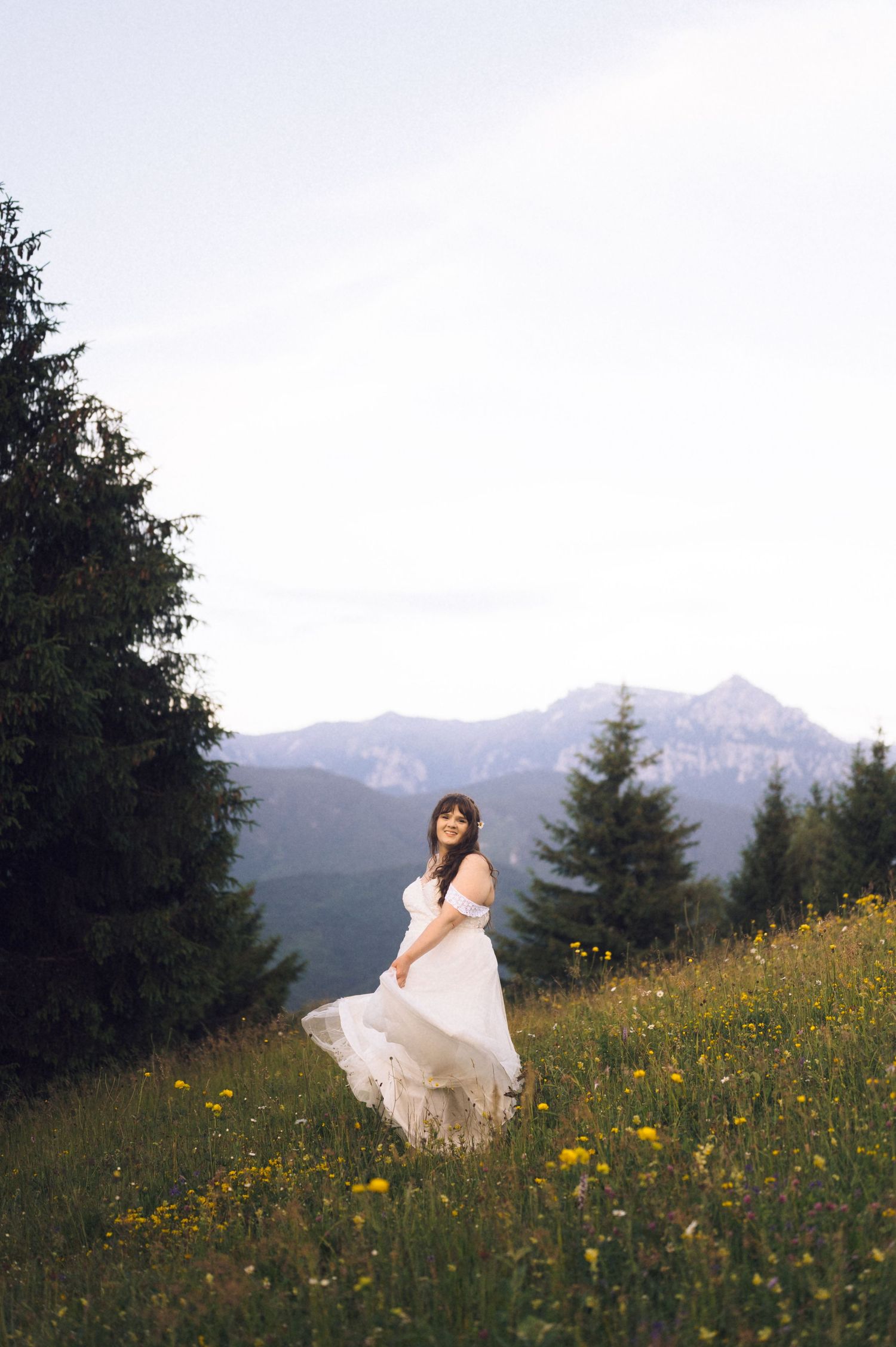 Bride in flowing white gown twirling in alpine meadow with wildflowers, evergreen trees, and mountain peaks in background.