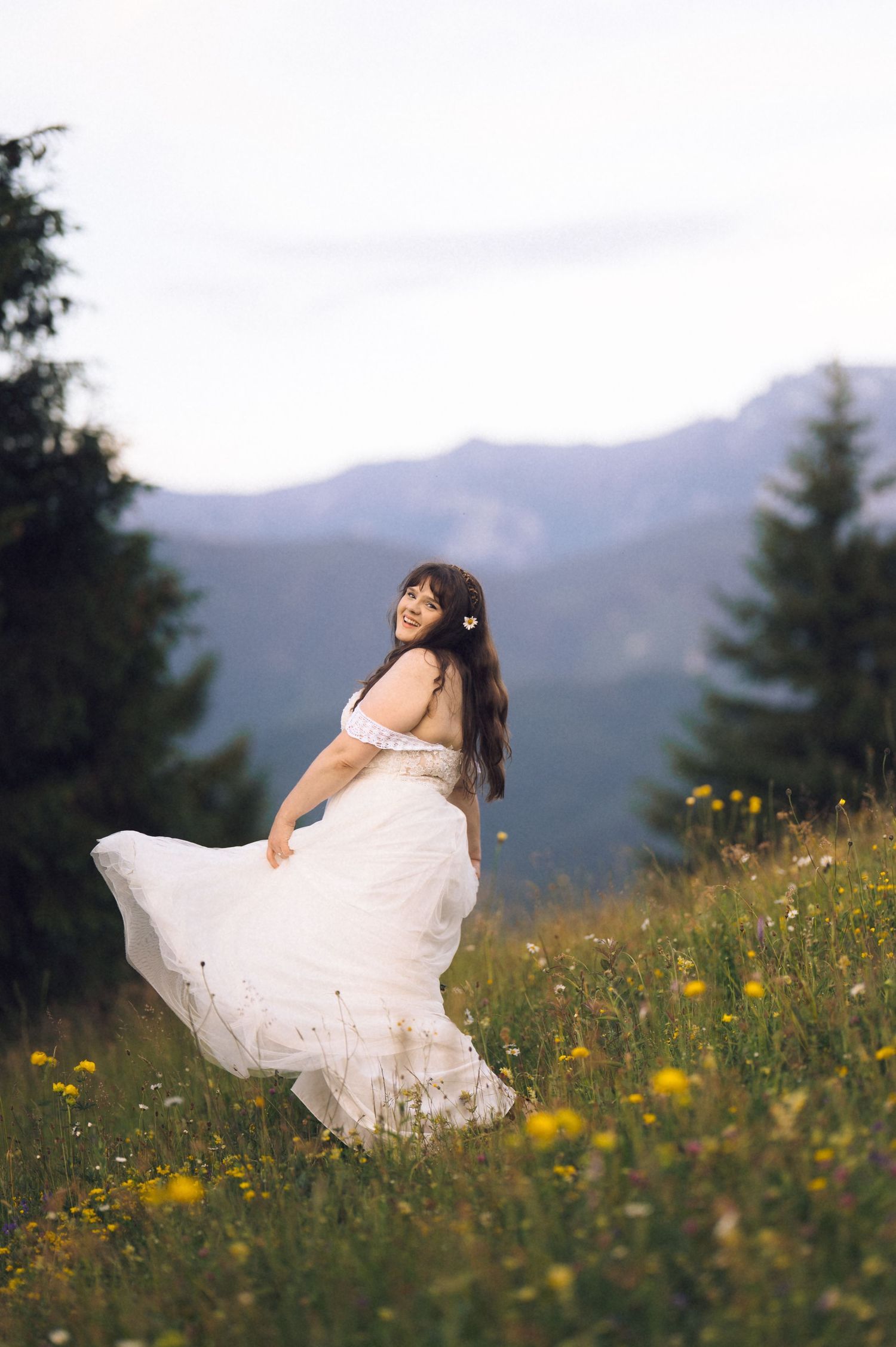 Smiling bride in flowing white dress twirling in a mountain meadow with wildflowers and evergreen trees.