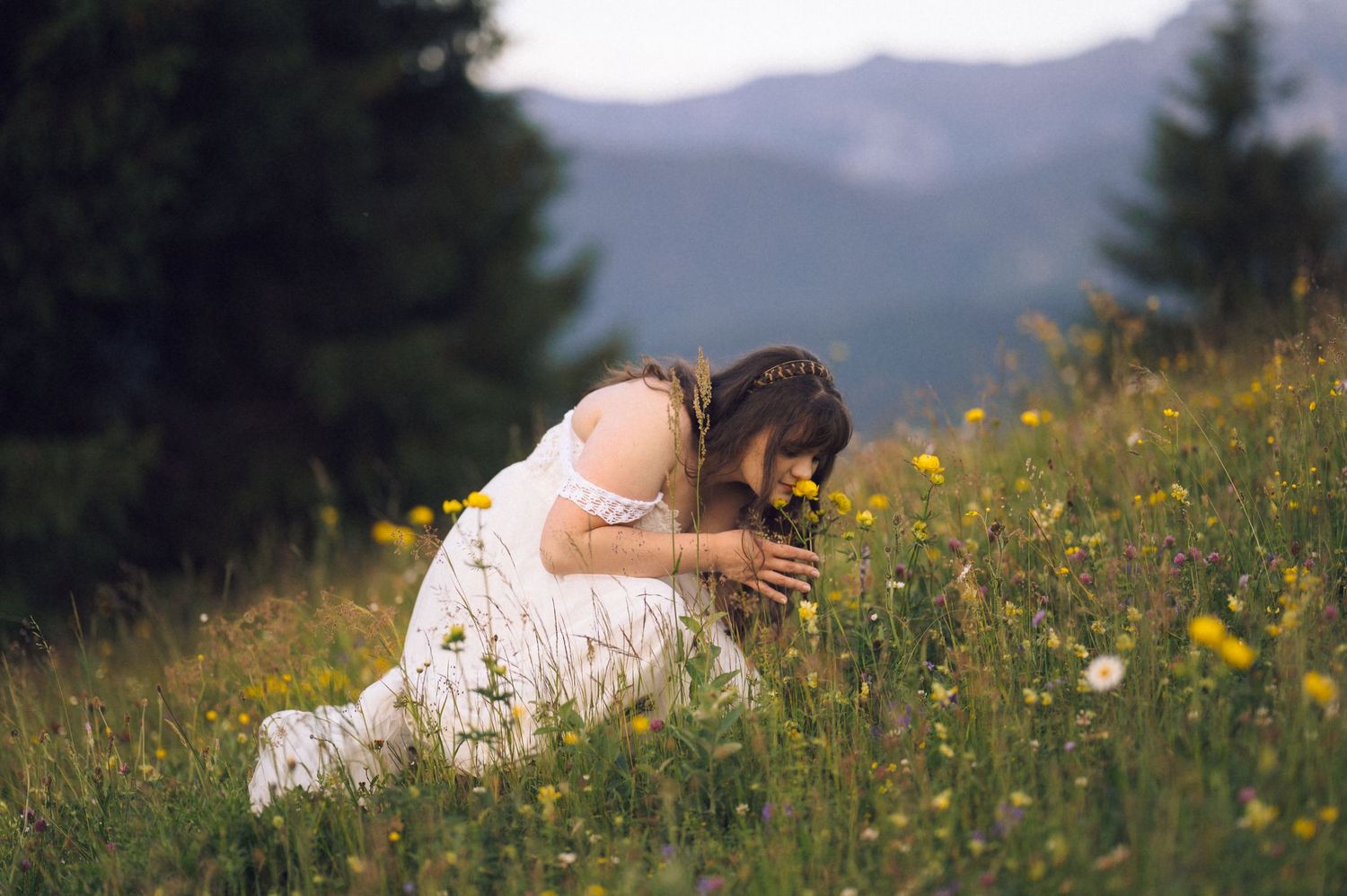 A young woman in a white dress kneels in a wildflower meadow, smelling yellow flowers with mountains in the background.